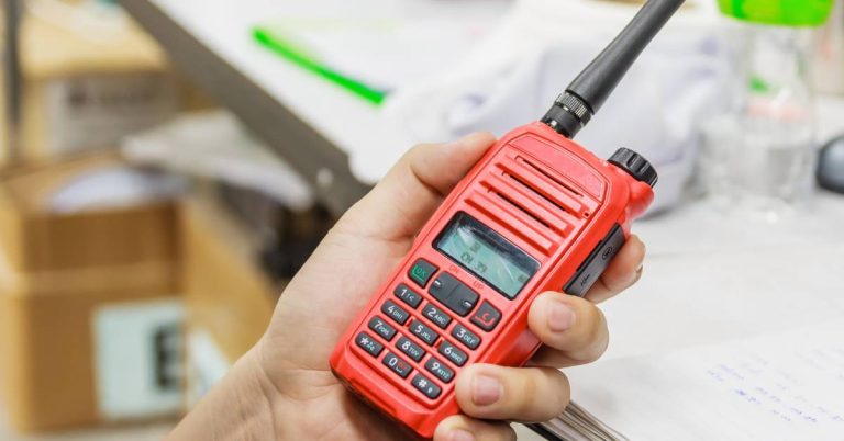 Close-up of a red two-way radio in a person's hand. In the background is a desk with stacks of paper on it.