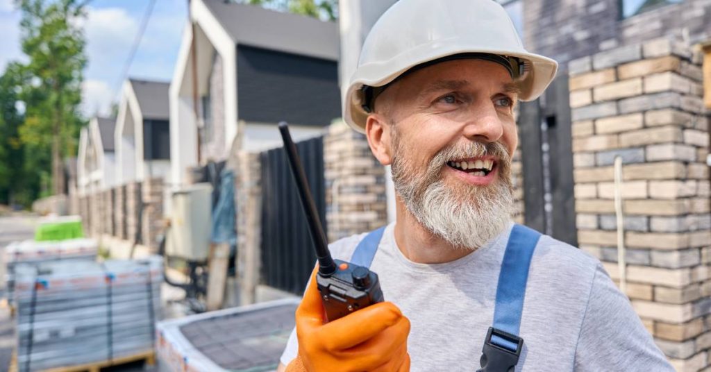 A man with a gray beard wearing blue overalls standing in front of pallets with a two-way radio in his hand.