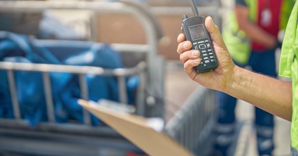 A person wearing high-visibility clothing while holding a clipboard in one hand and a two-way radio in the other.