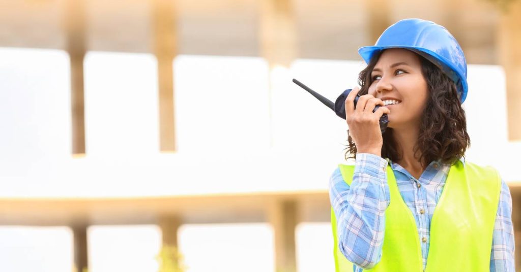 A woman wearing a blue hard hat and high-visibility work vest talking on a two-way radio while smiling.