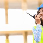 A woman wearing a blue hard hat and high-visibility work vest talking on a two-way radio while smiling.