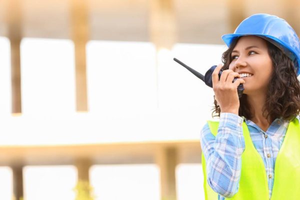 A woman wearing a blue hard hat and high-visibility work vest talking on a two-way radio while smiling.