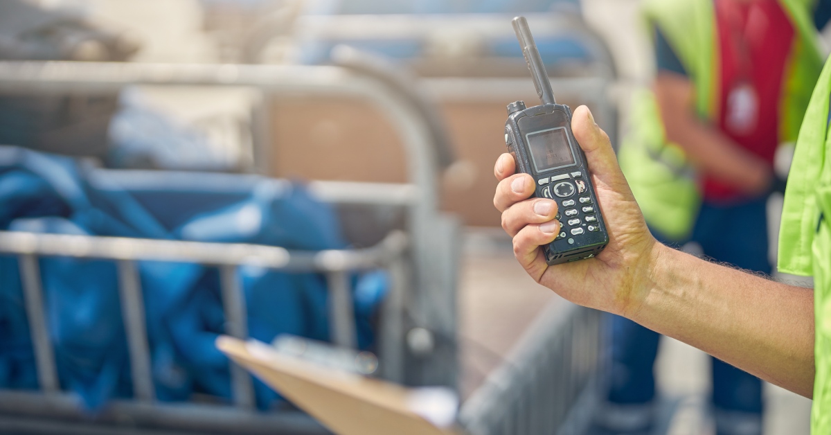 A person in a high-visibility shirt standing in a warehouse holding a clipboard in one hand and a two-way-radio in the other.