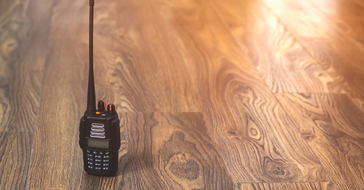 A black two-way radio with a blank screen, white buttons, and orange tabs on the knobs sitting on a hardwood floor.