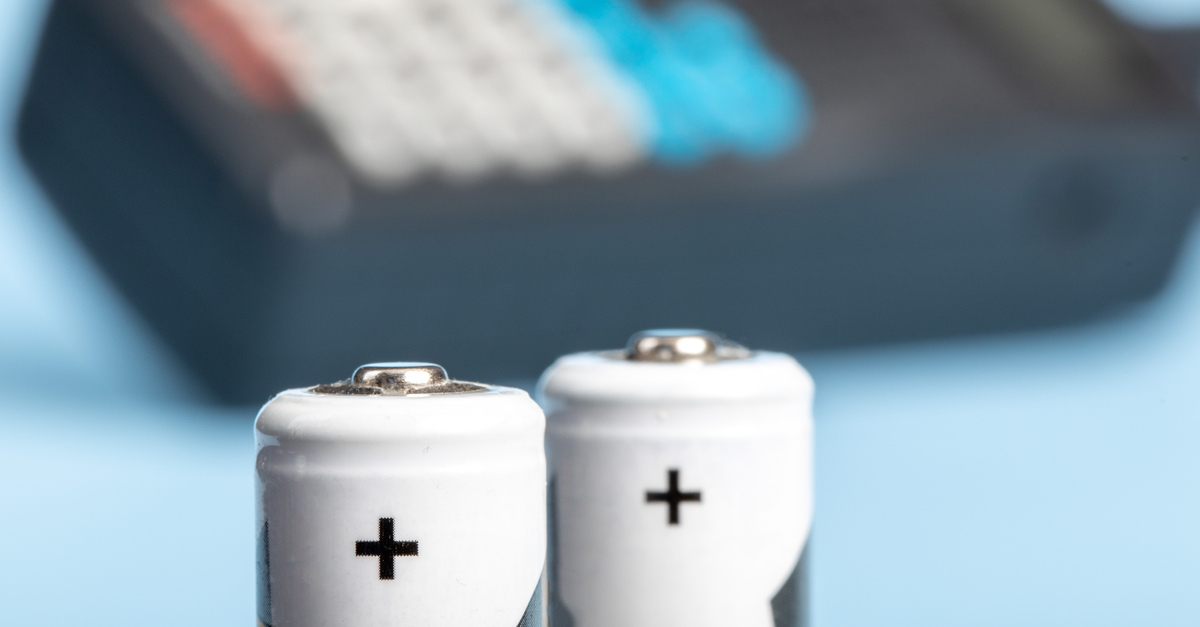 Two white batteries with the positive ends up in front of a blurry black radio in the background with blue buttons.