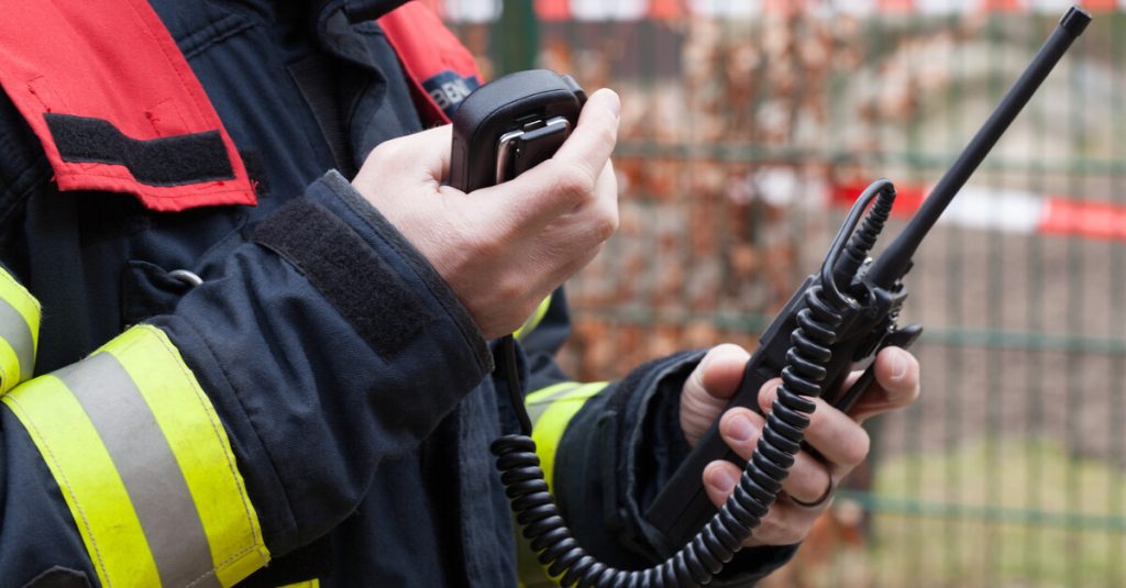 A firefighter wearing protective gear next to caution tape looking down at the two-way radio in his hand.