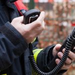 A firefighter wearing protective gear next to caution tape looking down at the two-way radio in his hand.