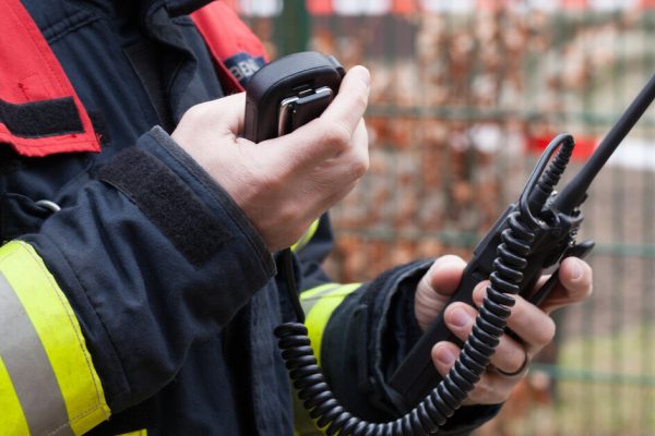 A firefighter wearing protective gear next to caution tape looking down at the two-way radio in his hand.