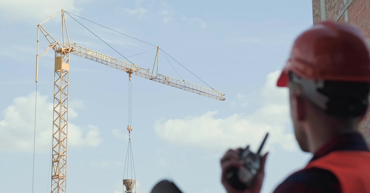 A man wearing a red hard hat and high-visibility safety vest talking on a two-way radio while looking at a crane.