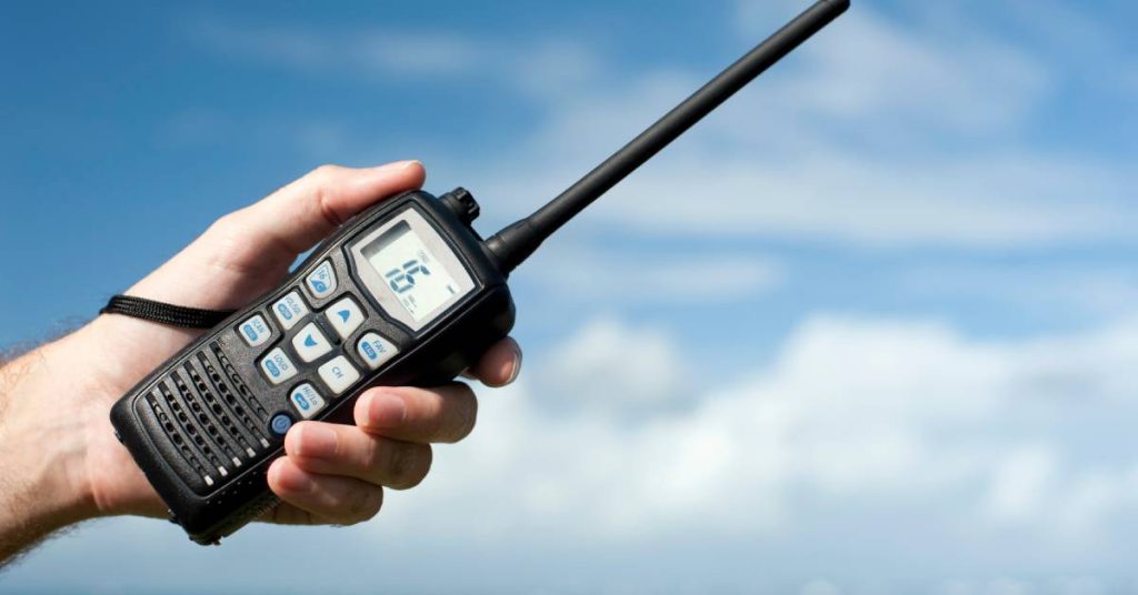 A person’s hand holding a black two-way radio with a strap on it. The screen shows the number "16".