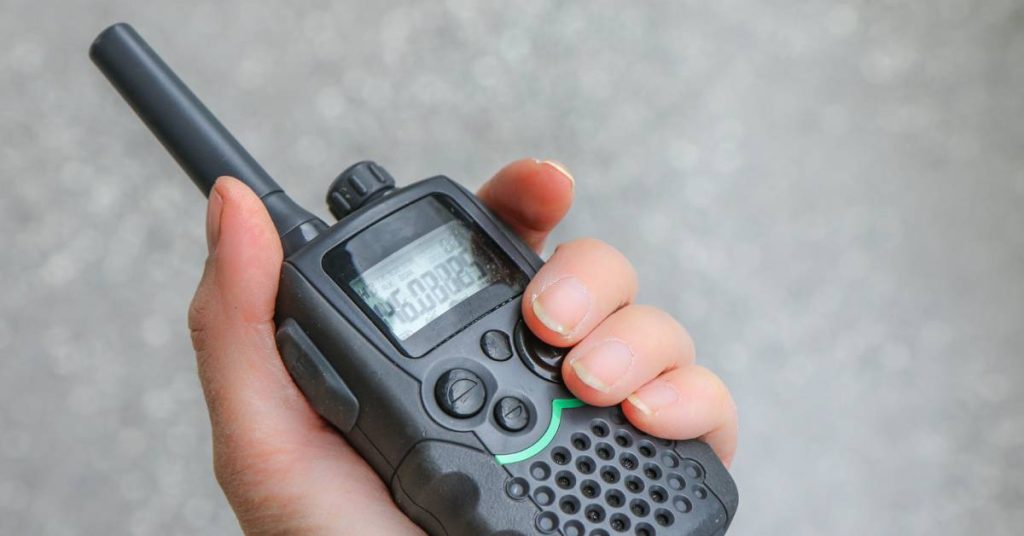 A close-up of a hand holding a black two-way radio with a green stripe on it. There are numbers displayed on the screen.