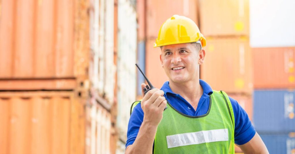 A man in a yellow hard hat and green high-visibility vest holding a two-way radio in his hand with a smile on his face.