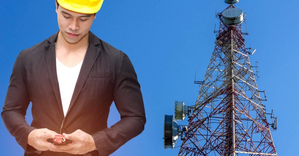 A man in a suit and yellow hard hat looking down at a two-way radio. There is a communications tower behind him.