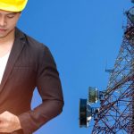 A man in a suit and yellow hard hat looking down at a two-way radio. There is a communications tower behind him.