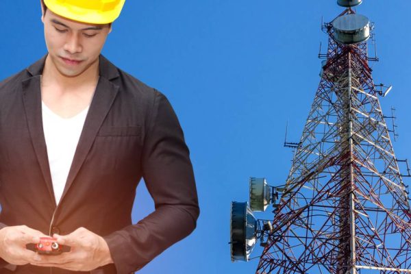 A man in a suit and yellow hard hat looking down at a two-way radio. There is a communications tower behind him.