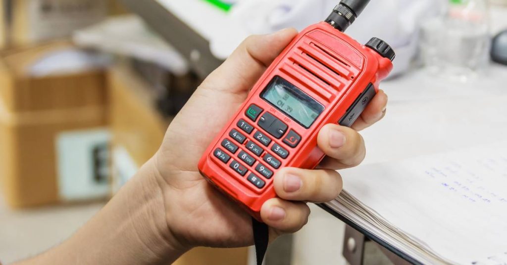 A person’s hand holding a red two-way radio with black buttons and dial. There’s a desk in the background.