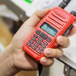 A person’s hand holding a red two-way radio with black buttons and dial. There’s a desk in the background.