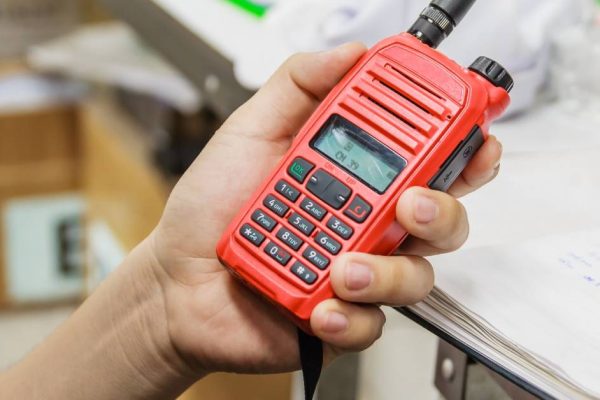 A person’s hand holding a red two-way radio with black buttons and dial. There’s a desk in the background.