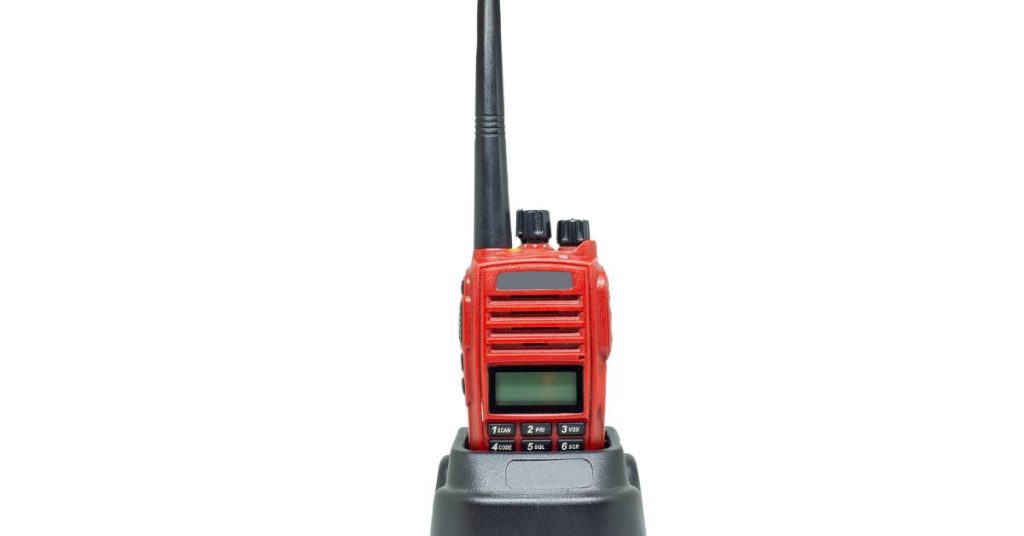 A red two-way radio with black buttons and antennae connected to a charging dock over a white background.