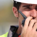 A construction worker wearing a white hardhat and a high-visibility vest talking into a radio in his hand.