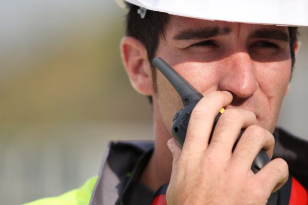 A construction worker wearing a white hardhat and a high-visibility vest talking into a radio in his hand.