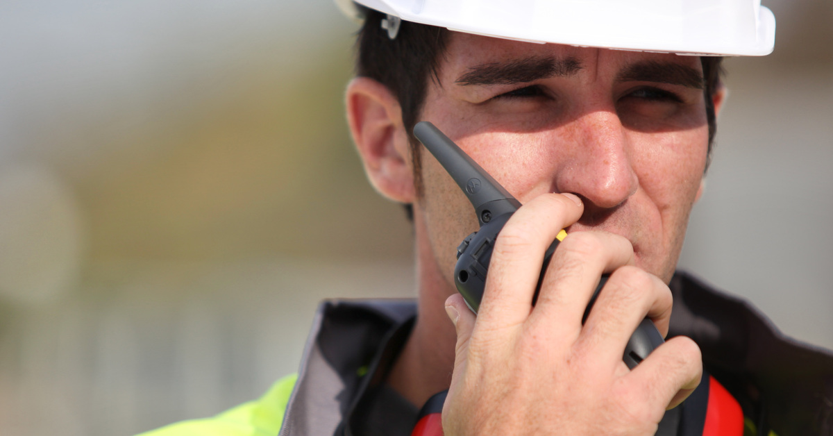 A construction worker wearing a white hardhat and a high-visibility vest talking into a radio in his hand.