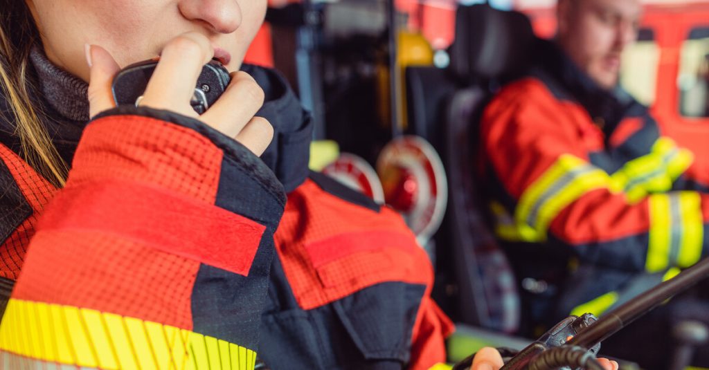 Two firefighters sitting in a truck wearing protective gear. The woman in the foreground is talking on a two-way radio.