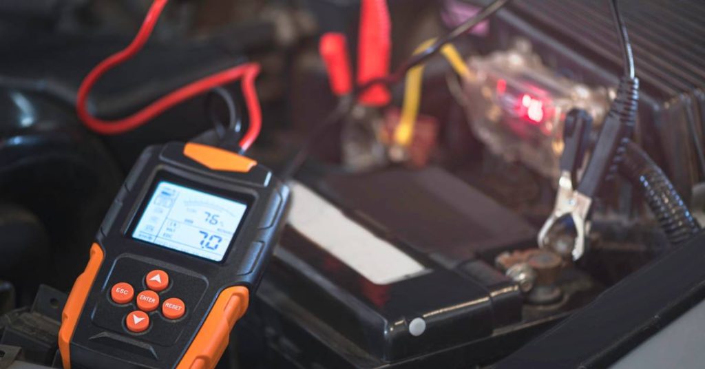 A digital battery tester sitting inside the engine bay of a car connected to the battery with readings on the screen.