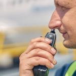 A close-up of a construction worker’s face in front of a blurry background. He is holding a two-way radio up to his mouth.