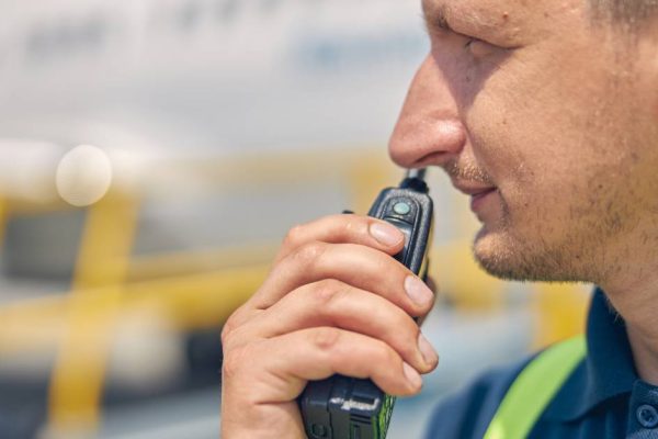 A close-up of a construction worker’s face in front of a blurry background. He is holding a two-way radio up to his mouth.