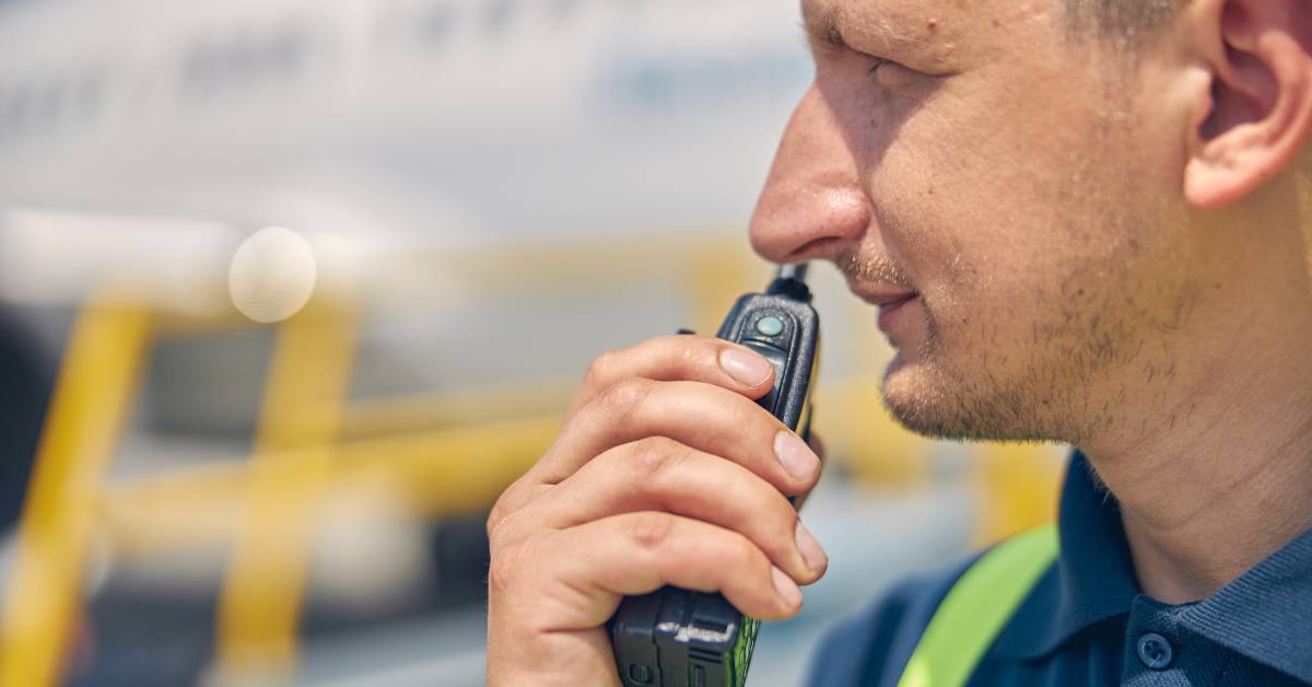 A close-up of a construction worker’s face in front of a blurry background. He is holding a two-way radio up to his mouth.