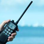 A person’s hand holding a two-way radio with 16 on the screen in front of the ocean with waves and clouds above.