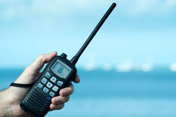 A person’s hand holding a two-way radio with 16 on the screen in front of the ocean with waves and clouds above.