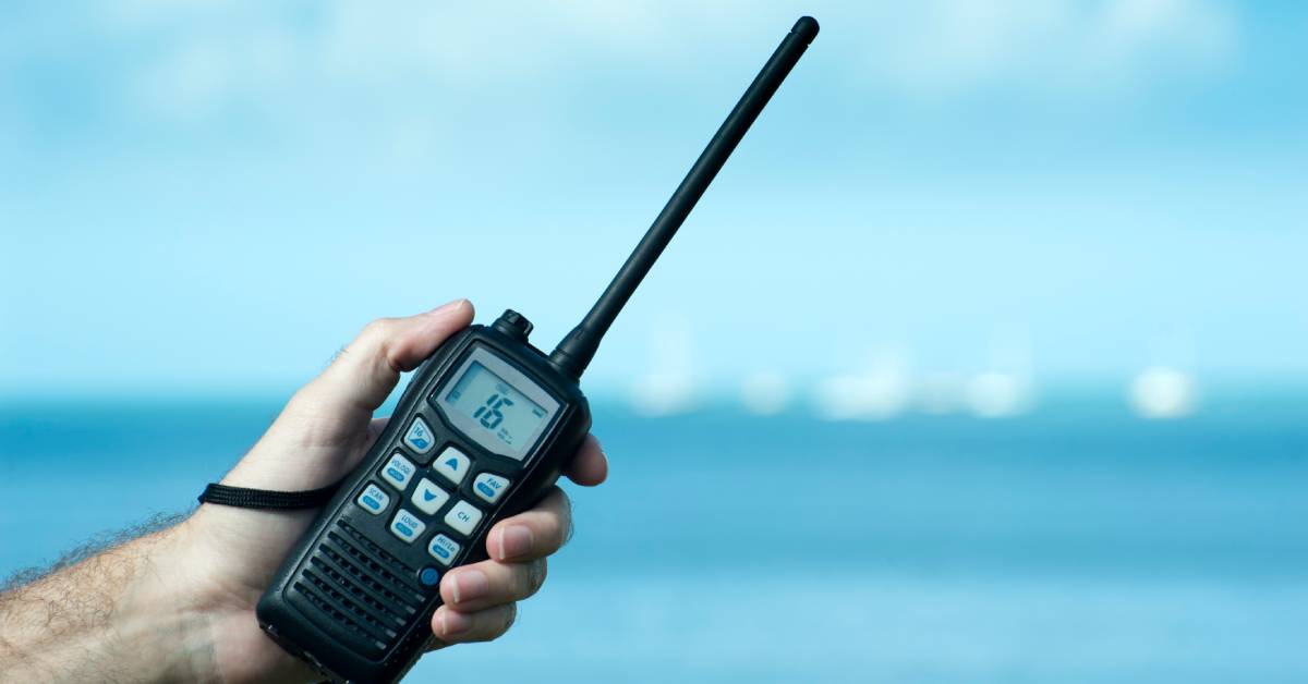 A person’s hand holding a two-way radio with 16 on the screen in front of the ocean with waves and clouds above.