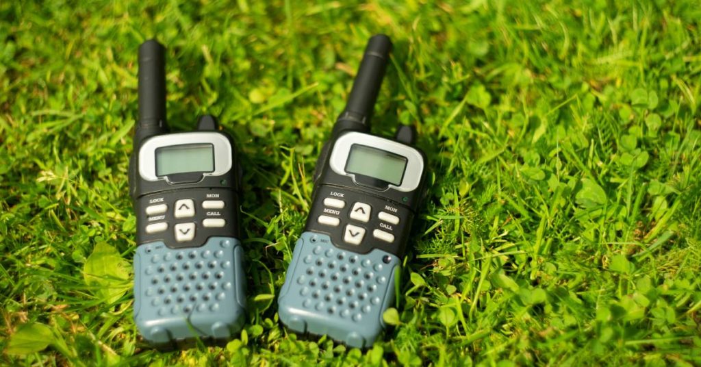 A pair of black and gray two-way radios sitting in the grass during the daytime with nothing on their displays.