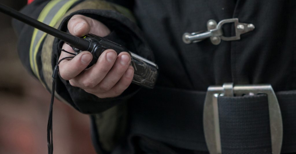 A person wearing black work gear with a red patch on the right shoulder holds a walkie-talkie radio in their hand.