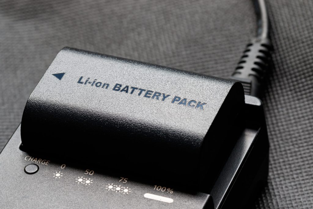 A black lithium-ion battery with a charger sits on a grey surface. The battery reads "Li-ion Battery Pack" in black letters.