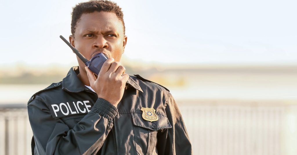 A Black police officer in a black uniform, with a gold badge and the word “Police” on his chest, talks into a radio.