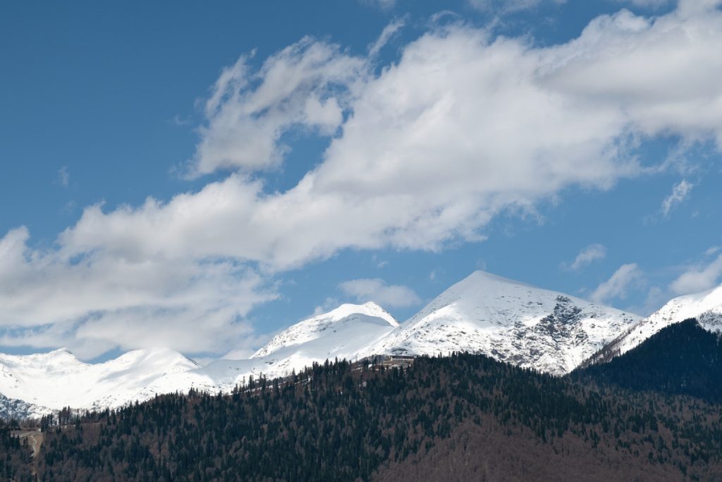 Snowy mountain peaks stand under white clouds and above smaller peaks with trees. The sun shines through the clouds.