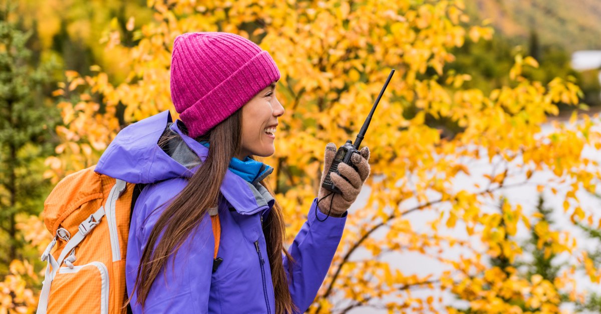 A female hiker wearing gloves, a hat, and an orange backpack while standing outside in autumn. She is using a two-way radio.