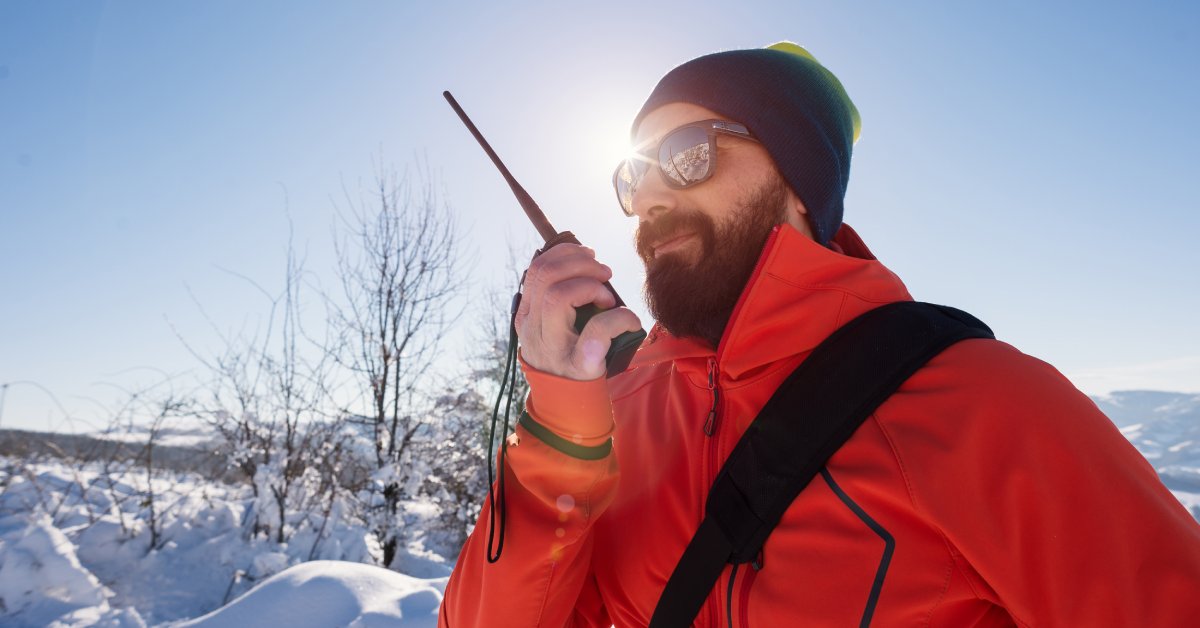 A man in a winter hat, red coat, and sunglasses holding a two-way radio while standing outside in the snow.