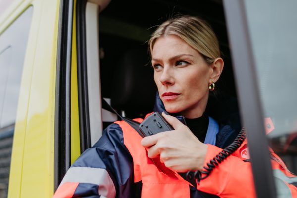 A blonde woman in an orange hi-vis jacket holding up a two-way radio while sitting in a red and yellow vehicle.