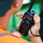 An over-the-shoulder view of a man in an orange shirt and black hat holding a black two-way radio in his right hand.