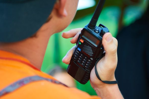 Hacks for Making Your Two-Way Radio Batteries Last Longer An over-the-shoulder view of a man in an orange shirt and black hat holding a black two-way radio in his right hand.
