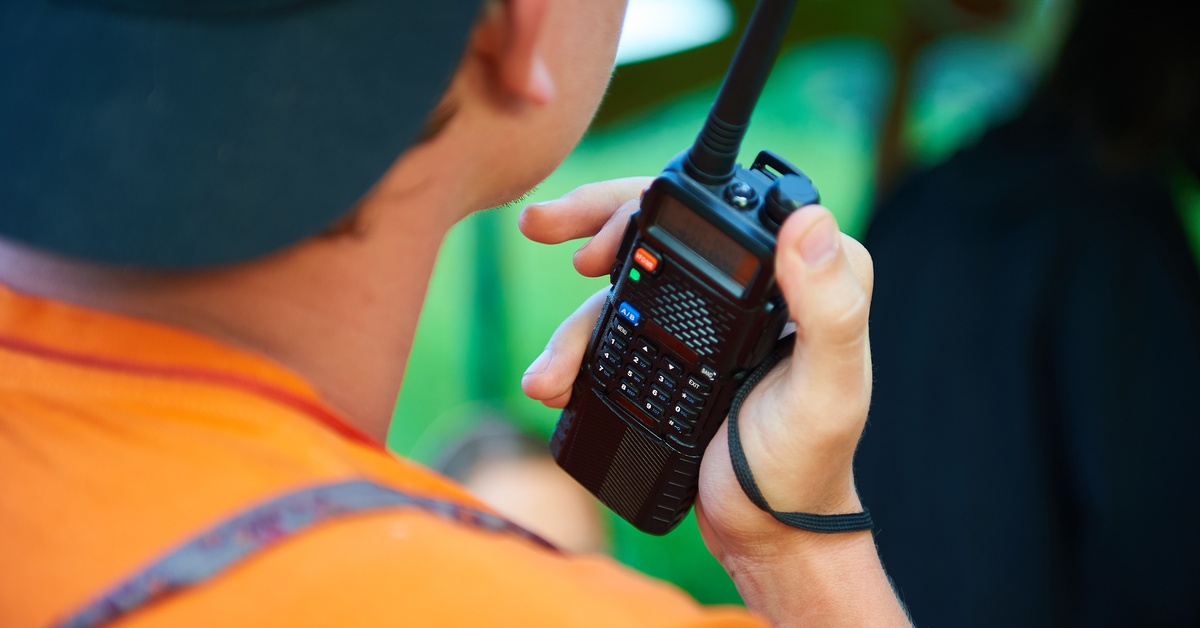 An over-the-shoulder view of a man in an orange shirt and black hat holding a black two-way radio in his right hand.