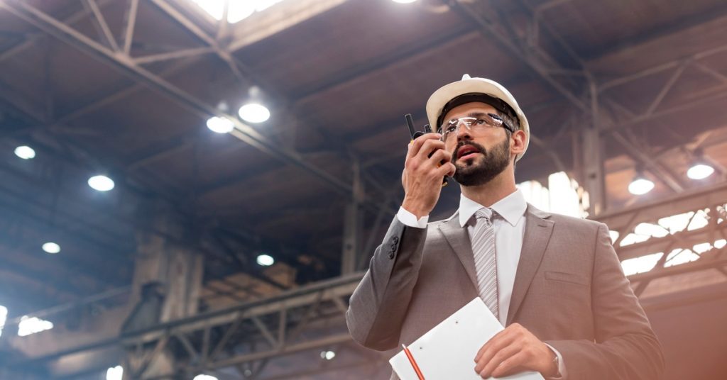 A man in a gray suit and white hard hat holding paperwork and a two-way radio while standing in a warehouse.