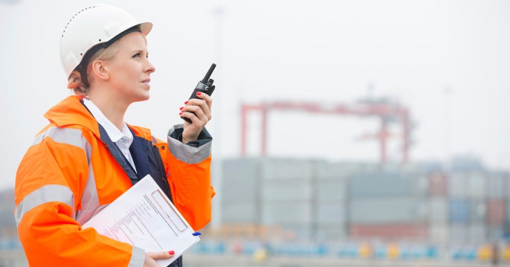 A woman in an orange coat and white hard hat holding paperwork and a two-way radio while standing in a shipping yard.