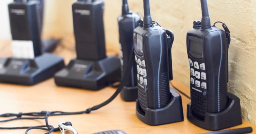 Several black two-way radios with white buttons and digital displays sitting in charging docks on a wooden table.