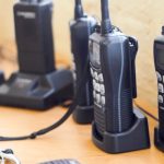 Several black two-way radios with white buttons and digital displays sitting in charging docks on a wooden table.
