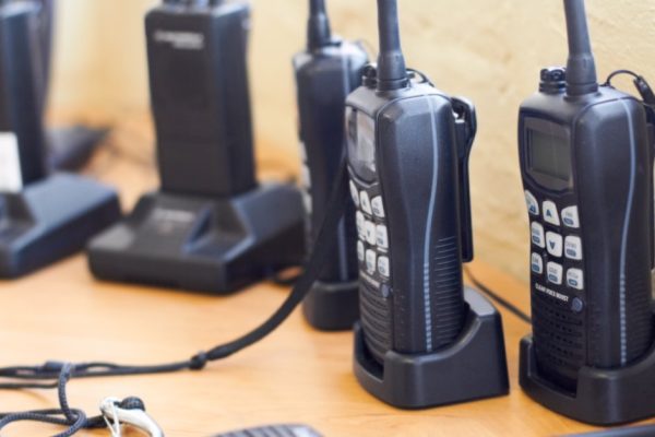 Several black two-way radios with white buttons and digital displays sitting in charging docks on a wooden table.